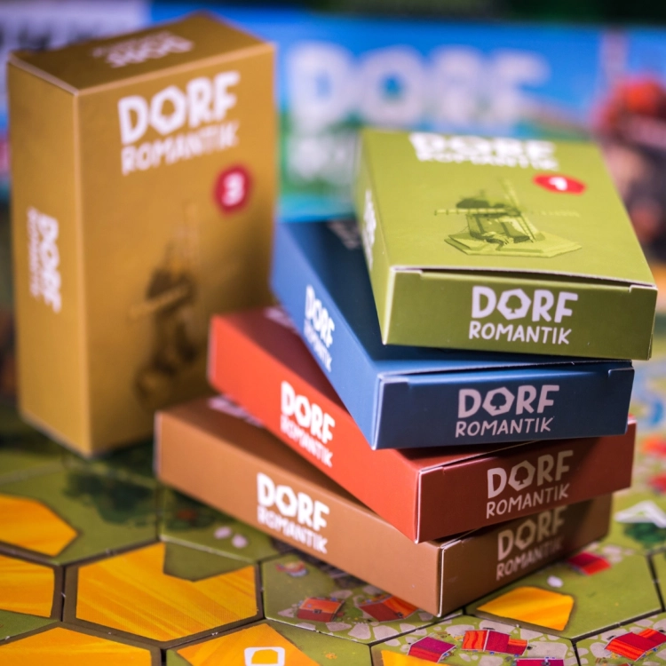 Stosy pudełek z elementami gry Dorfromantik – Kolorowe opakowania zawierające dodatkowe elementy gry.