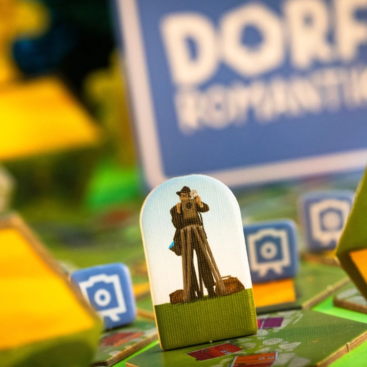 Figurka fotografa w grze Dorfromantik – Figurka fotografa na tle kolorowej planszy z napisem Dorfromantik.