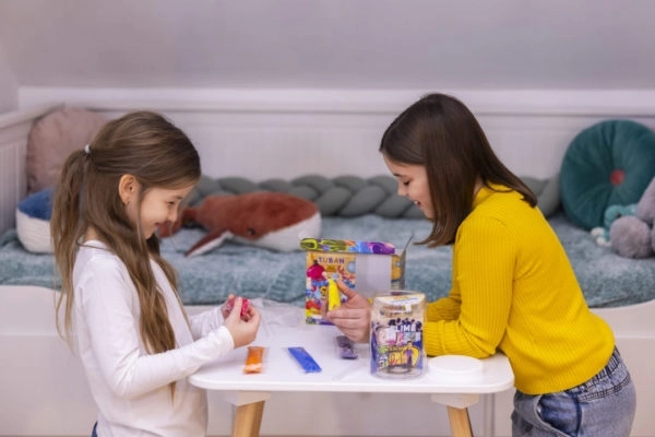 Dwie dziewczynki eksperymentujące z kolorowym piaskiem hydrofobowym przy stole w dziecięcym pokoju.