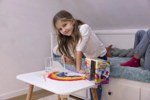 Dziewczynka tworząca kolorową kompozycję z piasku hydrofobowego na białym stole.