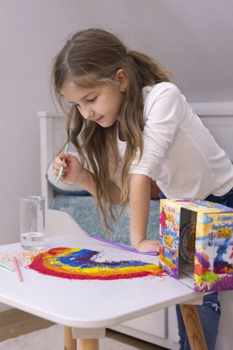 Zbliżenie na dziecko sypiące piasek hydrofobowy w formie tęczowego wzoru na kartce.