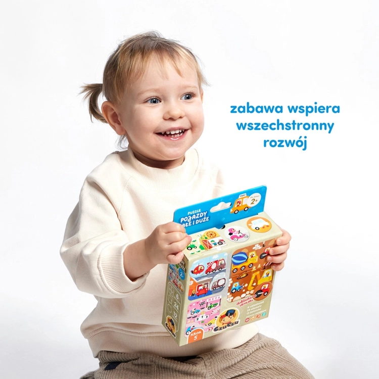 Dziecko uśmiechnięte trzyma pudełko z zestawem Puzzle Pojazdy małe i duże od CzuCzu.