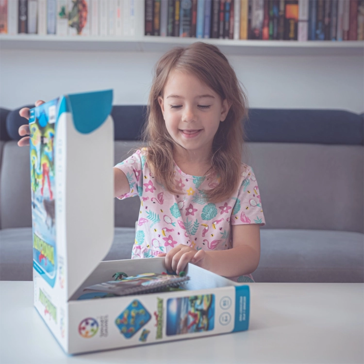 Dziewczynka z uśmiechem otwiera pudełko z grą Dinozaury – Tajemnicza Wyspa. W tle regał z książkami, a dziecko siedzi przy stole gotowe do zabawy.