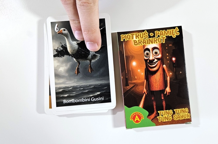 Pudełko gry Piotruś Brainrot Tung Sahur obok stosu kart, z górną kartą przedstawiającą Bombombini Gusini, trzymaną przez dłoń.