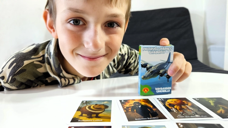 Młody chłopiec uśmiecha się, trzymając pudełko gry "Piotruś Pamięć Brainrot Bombardiro Crocodillo", obok rozłożonych kart z gry.
