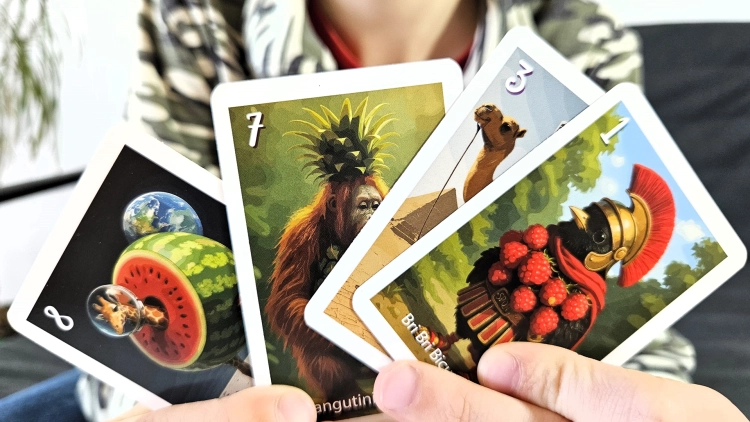 Dwie ręce trzymające cztery karty z gry "Piotruś Brainrot Bombardiro Crocodillo", ukazujące surrealistyczne ilustracje, takie jak arbuz-planeta i orangutan-ananas.