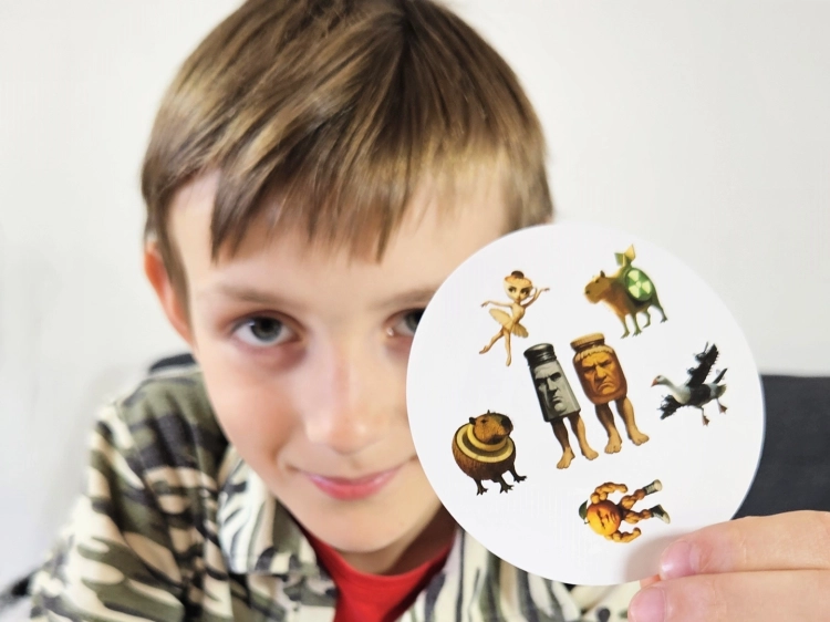 Chłopiec uśmiecha się, trzymając okrągłą kartę z gry "Brainrot Ale Pary" z różnorodnymi, dziwnymi postaciami.