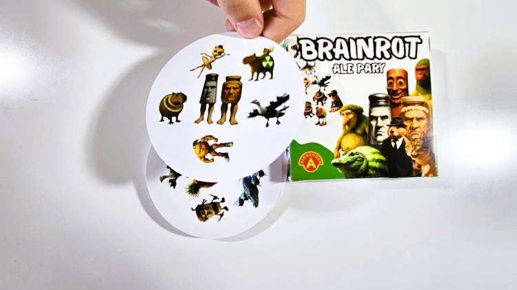 Ręka trzymająca dwie okrągłe karty z gry "Brainrot Ale Pary", z pudełkiem w tle.