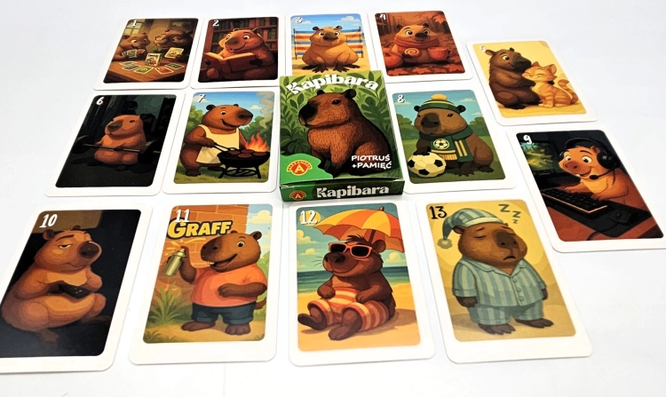 Pudełko gry "Piotruś Pamięć Kapibara" w centrum, otoczone rozłożonymi kartami z ilustracjami kapibar w różnych scenkach.