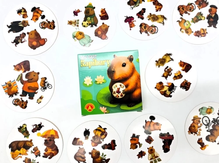 Pudełko gry "Ale Pary Kapibary" otoczone rozłożonymi okrągłymi kartami z ilustracjami kapibar.