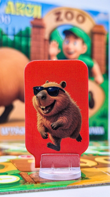 Zblliżenie na pionek kapibary w okularach przeciwsłonecznych na czerwonym tle, z pudełkiem gry w tle.