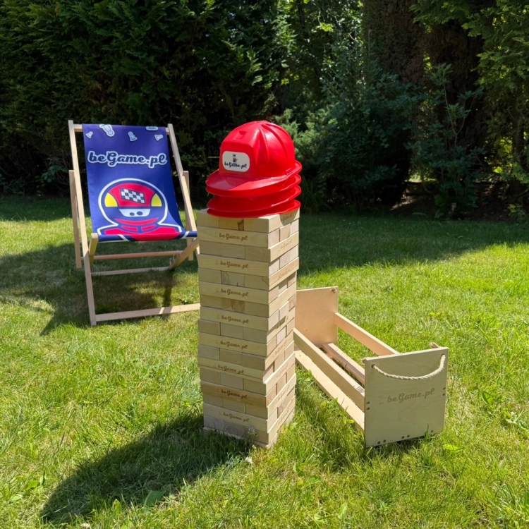 Wysoka wieża Jenga z naturalnych drewnianych klocków z logo BeGame.pl, stojąca na trawie obok leżaka i drewnianej skrzyni, z czerwonymi kaskami na szczycie.