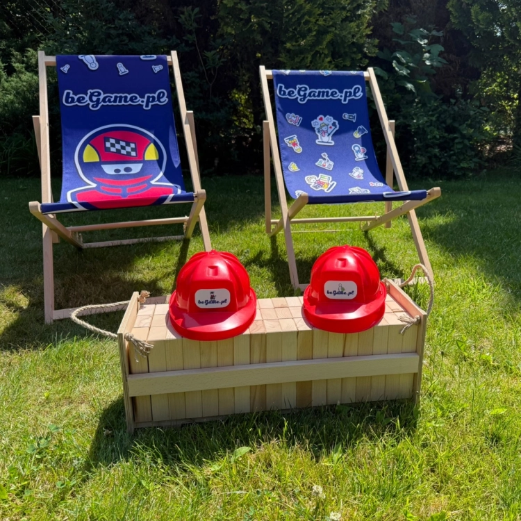 Dwa leżaki z logo BeGame.pl (jeden z grafiką kierowcy wyścigowego, drugi z ikonami gier) oraz drewniana skrzynia z klockami Jenga i dwoma czerwonymi kaskami na trawie.