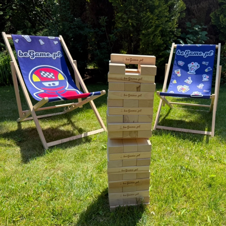Wysoka wieża Jenga z naturalnych drewnianych klocków z logo BeGame.pl, stojąca na trawie między dwoma leżakami z logo BeGame.pl.