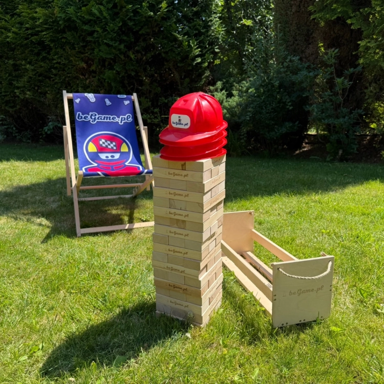 Wysoka wieża Jenga z naturalnych drewnianych klocków z logo BeGame.pl, stojąca na trawie obok leżaka i drewnianej skrzyni, z czerwonymi kaskami na szczycie.