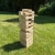Wysoka wieża Jenga zbudowana z naturalnych drewnianych klocków z logo BeGame.pl, stojąca na zielonej trawie, sprawiająca wrażenie chwiejnej.