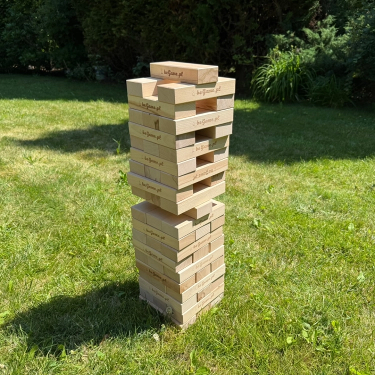 Wysoka wieża Jenga zbudowana z naturalnych drewnianych klocków z logo BeGame.pl, stojąca na zielonej trawie, sprawiająca wrażenie chwiejnej.
