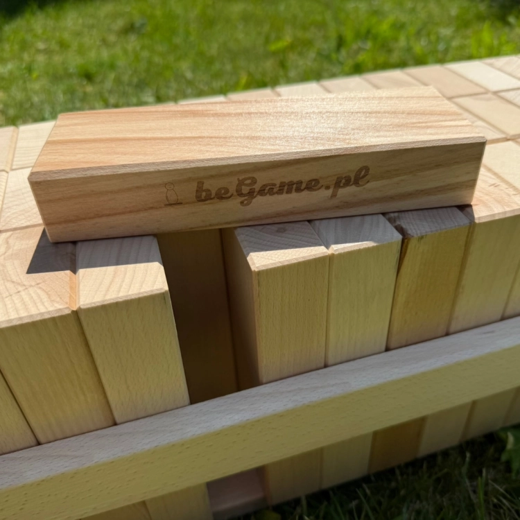 Zblliżenie na pojedynczy drewniany klocek z logo "beGame.pl" leżący na szczycie dużej wieży Jenga, widocznej w tle na trawie.