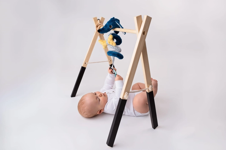 Niemowlę leży na plecach pod drewnianym pałąkiem, łapiąc zawieszoną granatową spiralę edukacyjną Jamiś Mom's Care w kształcie psa. Wzory kontrastowe.