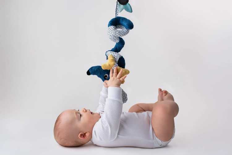 Niemowlę na plecach, podciąga się do wiszącej spirali edukacyjnej Jamiś Mom's Care. Dziecko leży na białym tle, wyciąga rączki do zabawki.