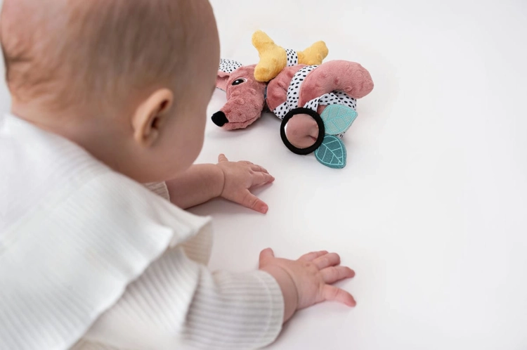 Niemowlę w białym ubranku patrzy i sięga rączkami do różowej spirali edukacyjnej Jamiś z czarno-białymi wzorami i żółtymi elementami, leżącej na białym tle.