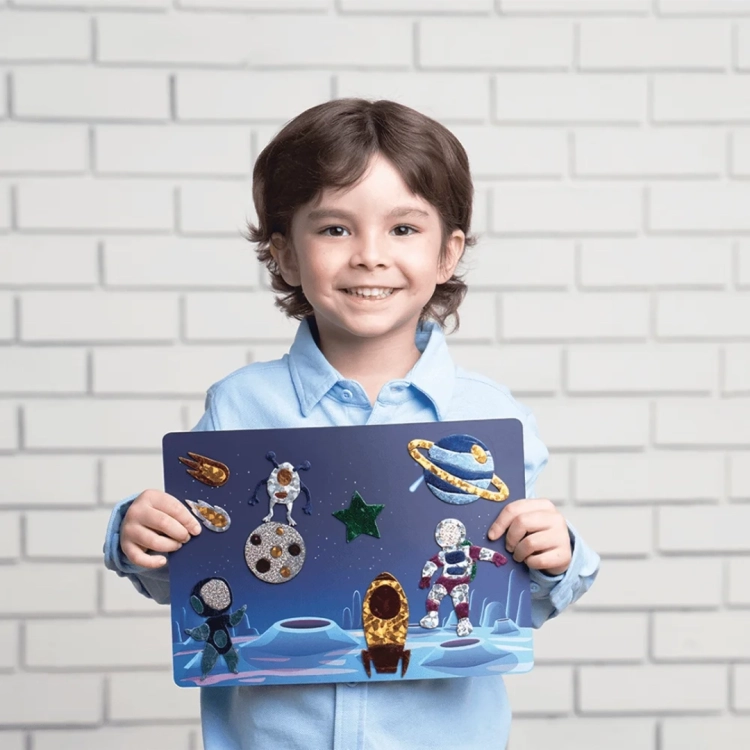 Chłopiec uśmiecha się, trzymając gotowy obrazek z kosmitami i rakietami z zestawu Foil Fun.