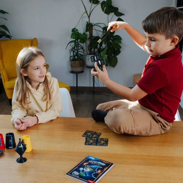 Chłopiec wykonuje sztuczkę magiczną z różdżką i kartami, siedząc na stole.