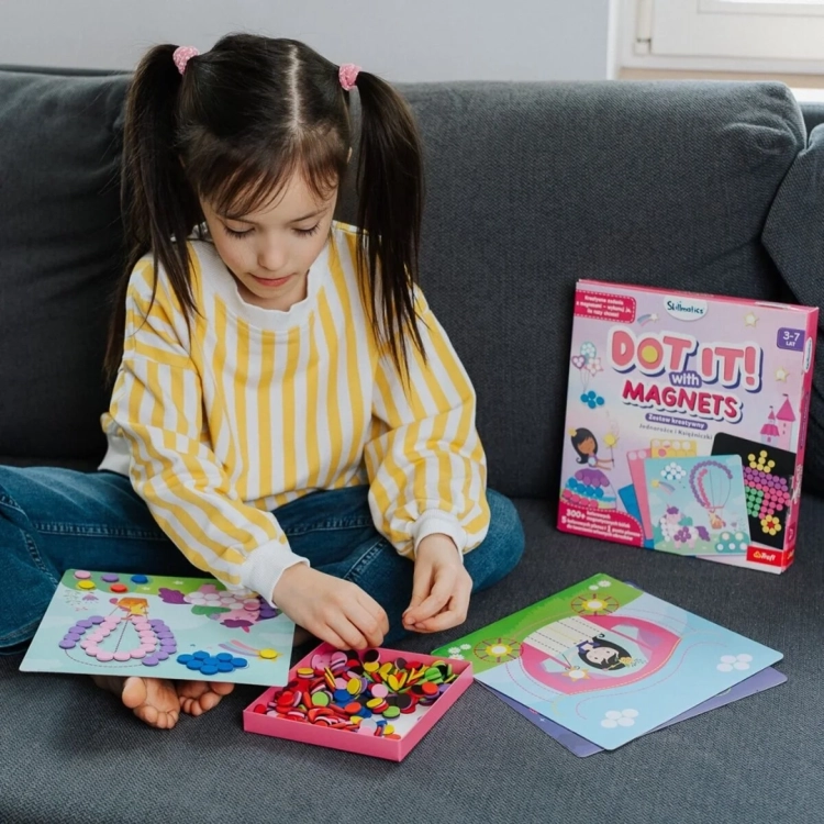 Dziewczynka w żółtym swetrze bawi się zestawem Skillmatics Dot It! z magnesami na sofie, układając kółka na planszy z Księżniczką i Jednorożcem.