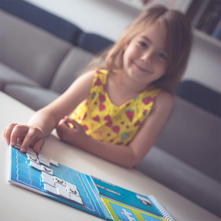 Uśmiechnięta dziewczynka układa elementy magnetyczne Parada Pingwinów Smart Games na niebieskiej planszy.