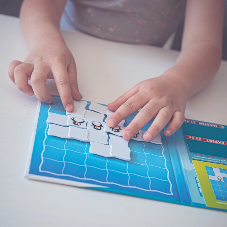 Dziecięce dłonie układają na planszy magnetyczne elementy z pingwinami z gry logicznej Parada Pingwinów Smart Games.