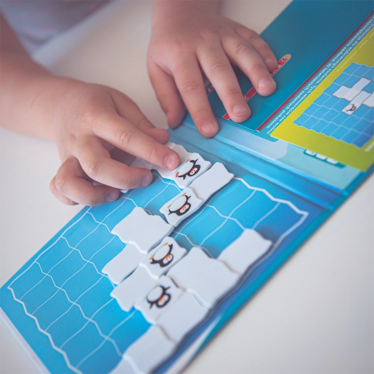 Zbliżenie na dłonie dziecka układającego elementy z pingwinami na planszy magnetycznej Parada Pingwinów Smart Games.