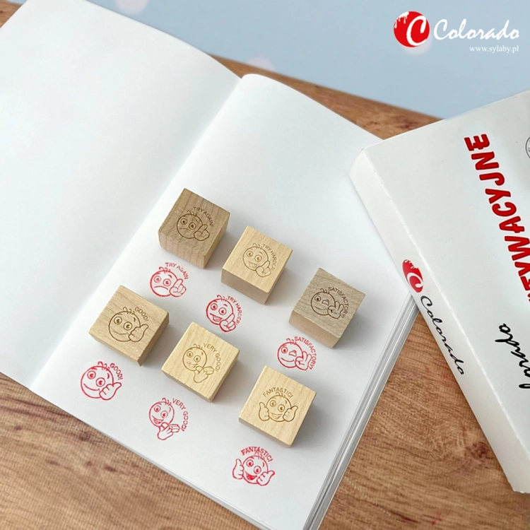 Zestaw 6 sześciennych drewnianych stempli motywacyjnych ANGIELSKIE Colorado 3+ na otwartym zeszycie z odbitymi emotikonami i ocenami (Fantastic, Good, Very Good, Try Again, Satisfactory)