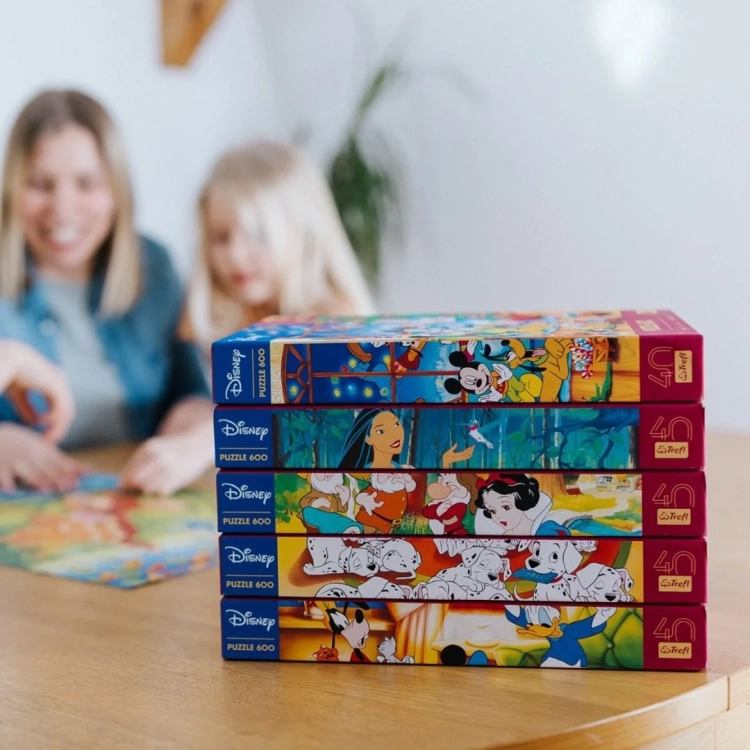 Stos pudełek z puzzlami Trefl Disney 600 elementów, w tle dorosła kobieta i dziecko układające puzzle.