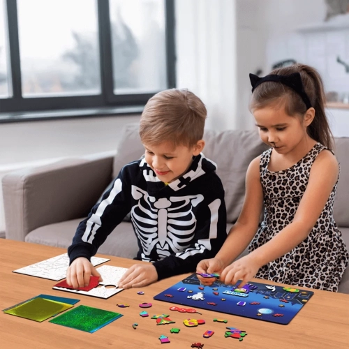 Dwoje dzieci w przebraniach halloweenowych siedzi przy stole, wyklejając kolorową folią piankowe naklejki i umieszczając je na planszy z nocnym krajobrazem.