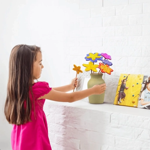 Dziewczynka w różowej sukience wkłada samodzielnie ozdobiony, kolorowy kwiatek do wazonu stojącego na białej półce.