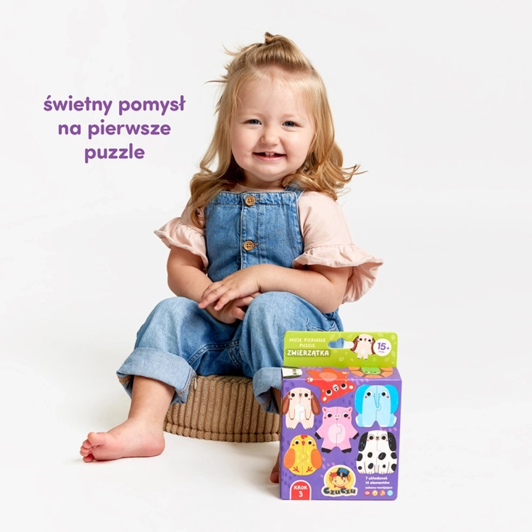 Mała dziewczynka w dżinsowych ogrodniczkach uśmiecha się, siedząc obok fioletowego pudełka z puzzlami CzuCzu.