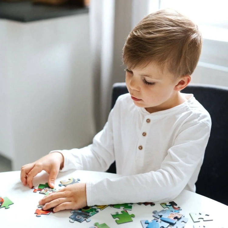 Chłopiec w białej bluzce siedzi przy stole i z koncentracją dopasowuje do siebie duże elementy puzzli.