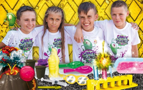 Czworo dzieci w koszulkach Crazy Science pozuje przy stanowisku z akcesoriami do przeprowadzania eksperymentów, takimi jak próbówki, strzykawka i makieta wulkanu.