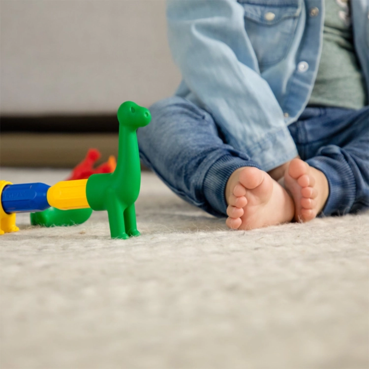 Zbliżenie na połączone magnetycznie elementy zielonego dinozaura stojącego na jasnym, puszystym dywanie obok stóp dziecka.