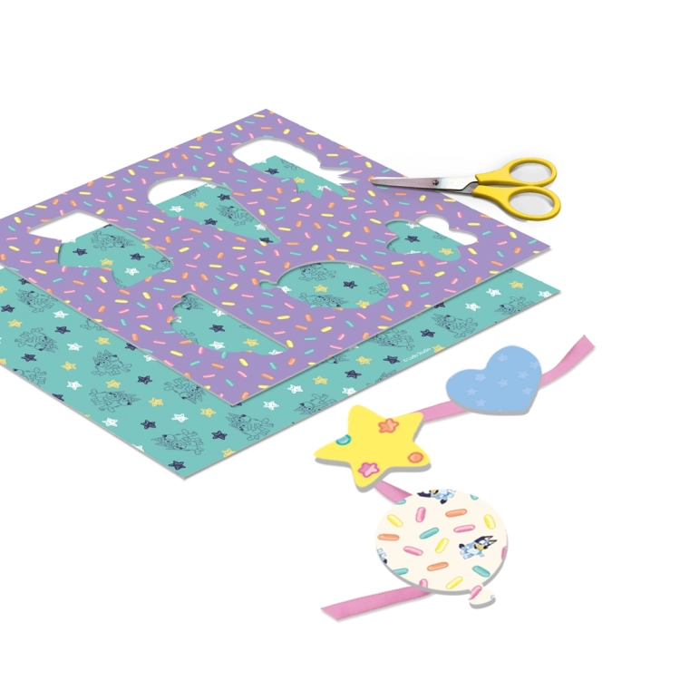 Kolorowe arkusze papieru z wzorami, fioletowy szablon z otworami, żółte nożyczki oraz ozdobna girlanda z papierowymi elementami.