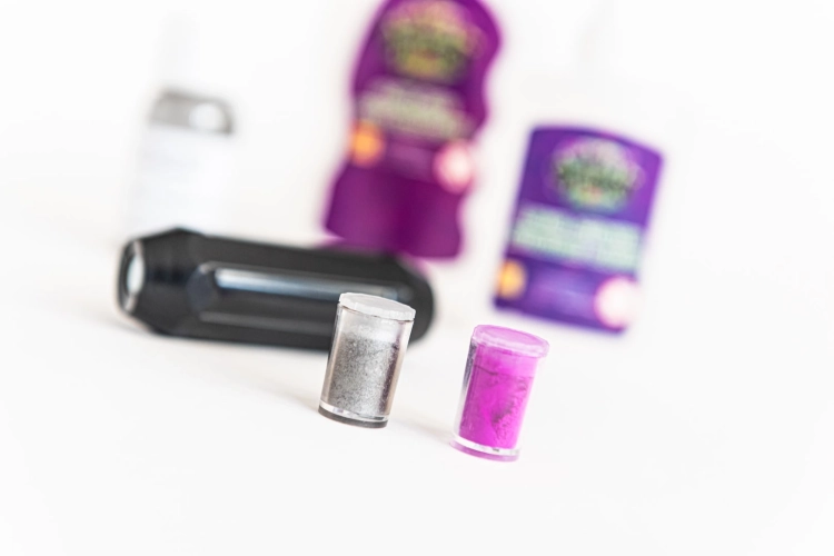 Zbliżenie na dwa małe pojemniki z sypkim pigmentem w kolorze srebrnym i fioletowym. W tle widać rozmyte butelki z klejem i aktywatorem do tworzenia kreatur.