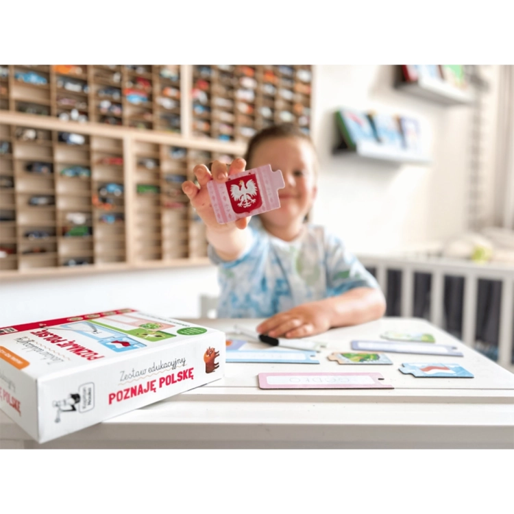 Uśmiechnięte dziecko prezentuje element puzzli z polskim godłem. Na stole leży pudełko zestawu i inne części układanki do pisania.