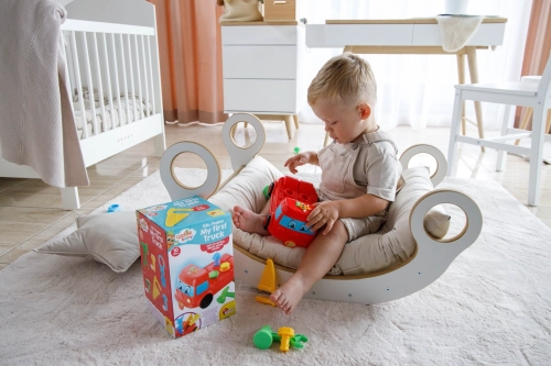 Boso siedzący na poduszce bujaka chłopiec wyjmuje z pudełka czerwoną ciężarówkę-sorter „Moje pierwsze auto” Lisciani. Wokół na dywanie leżą kolorowe elementy zestawu, w tym żółta piła i zielony młotek.