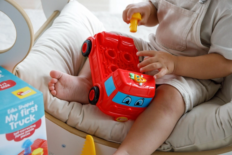 Czerwona ciężarówka-sorter Lisciani Carotina Baby z akcesoriami leżąca na drewnianym blacie obok otwartego pudełka. W tle widoczne okno z firanką, roślina w doniczce i otwarta książeczka dla dzieci.