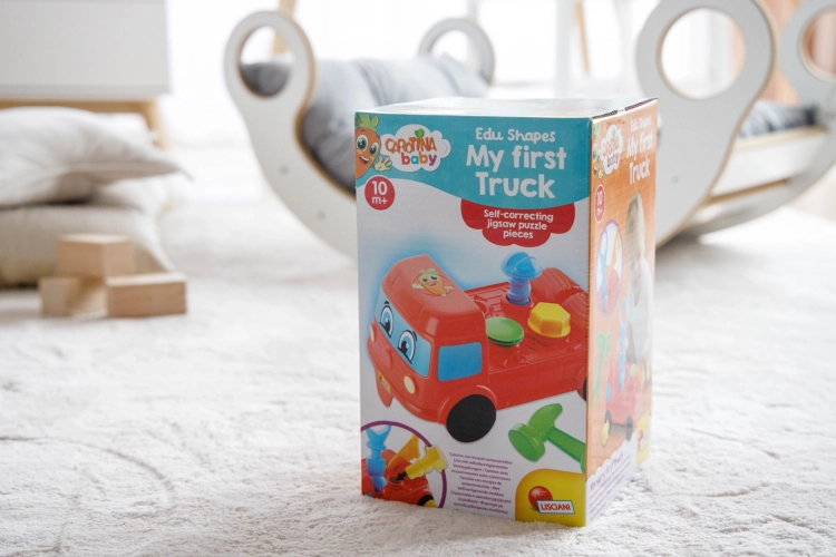 Pudełko z zabawką edukacyjną Lisciani Carotina Baby „Moje pierwsze auto” stoi na jasnym, puszystym dywanie. W tle widać fragment białego bujaka z poduszką oraz beżowe poduszki dekoracyjne.