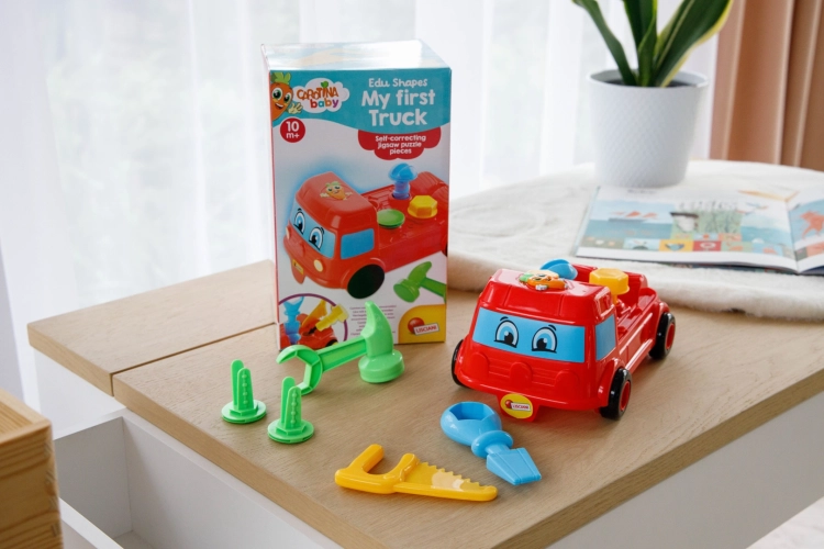 Czerwona ciężarówka-sorter Lisciani Carotina Baby z akcesoriami leżąca na drewnianym blacie obok otwartego pudełka. W tle widoczne okno z firanką, roślina w doniczce i otwarta książeczka dla dzieci.