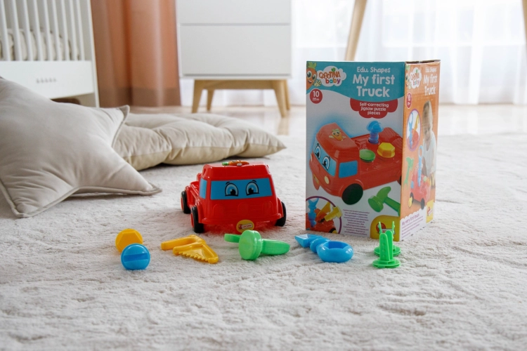 Czerwona ciężarówka Lisciani Carotina Baby z rozrzuconymi wokół kolorowymi akcesoriami (śruby, młotek, piła, klucz) na puszystym dywanie, obok kartonowego pudełka z zabawką. W tle łóżeczko.