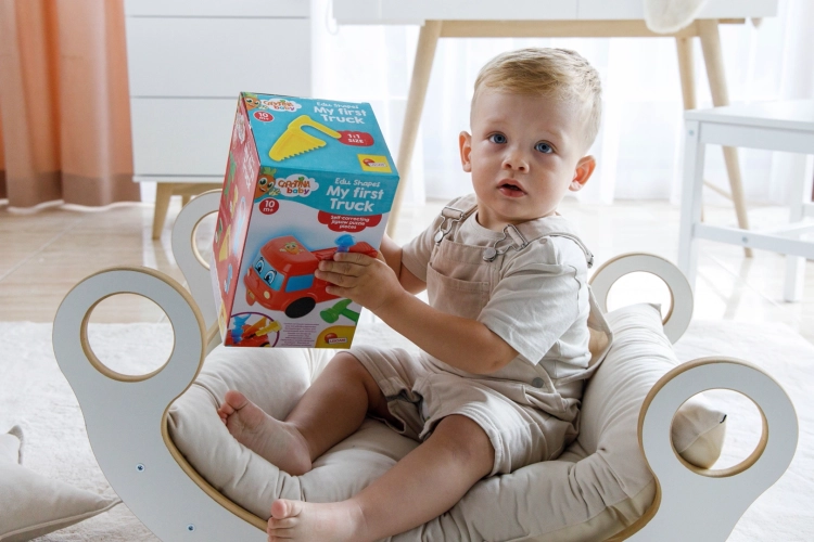 Chłopiec w beżowych ogrodniczkach trzyma w rękach kolorowe pudełko z zabawką edukacyjną „Moje pierwsze auto” Lisciani Carotina Baby. Siedzi boso na beżowej poduszce w białym, bujanym krzesełku na jasnym dywanie.