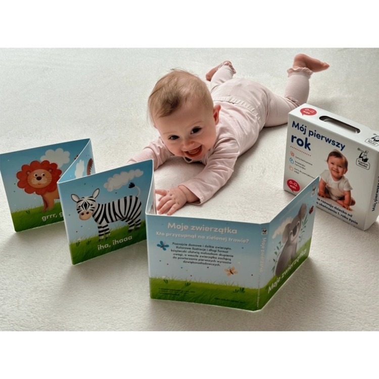 Uśmiechnięte niemowlę na brzuszku obok rozłożonej książeczki harmonijki „Moje zwierzątka”. W tle pudełko zestawu kontrastowego Kapitan Nauka.