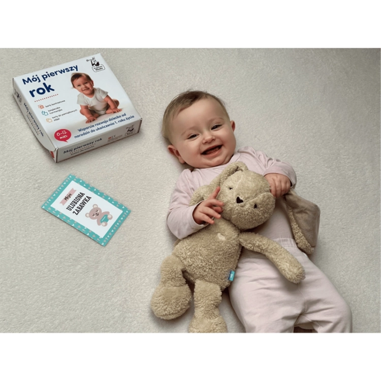 Radosne niemowlę leży na dywanie, przytulając pluszowego królika. Obok karta „Moja ulubiona zabawka” oraz opakowanie zestawu „Mój pierwszy rok”.